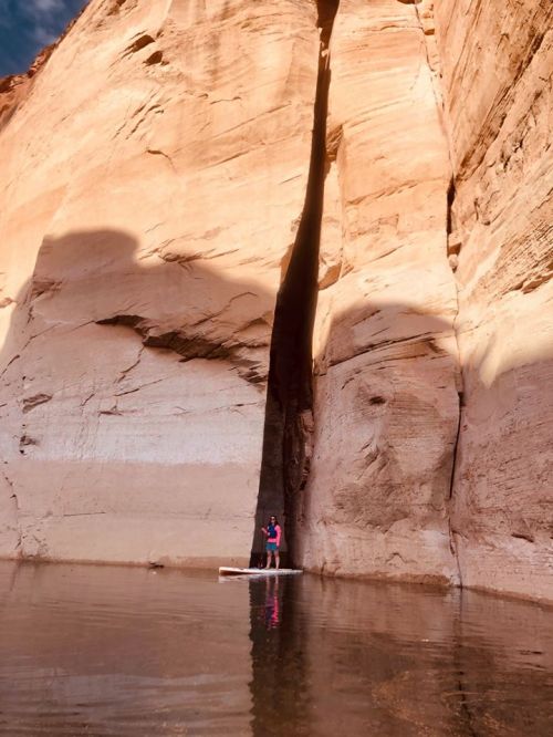 Two people stand on a raft in calm water, surrounded by towering, smooth rock walls in a narrow canyon.