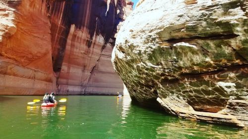 Two kayakers paddle through a narrow canyon with towering red rock walls and green water reflecting sunlight.