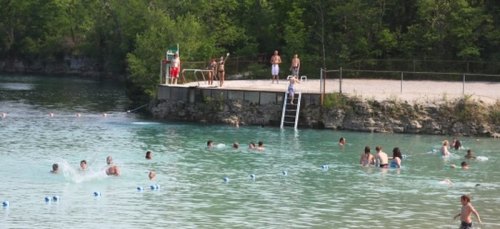 A sunny swimming area with people enjoying the water, a diving board, and a lifeguard on duty.