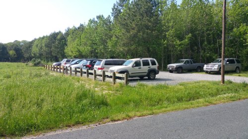 A gravel parking lot with several parked SUVs and a wooden fence, surrounded by green trees and grass.