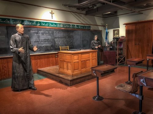 A historical classroom scene with life-sized figures of a priest and a nun, a wooden desk, and chalkboard.