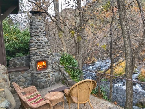 Cozy outdoor seating by a stone fireplace, overlooking a serene stream and autumn trees.