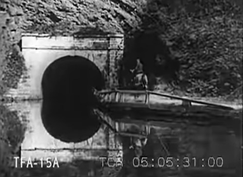 A person in a small boat approaches a dark tunnel entrance, surrounded by rocky terrain and calm water.
