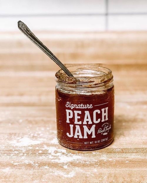 A jar of Signature Peach Jam with a spoon resting inside, placed on a wooden surface with flour scattered around.