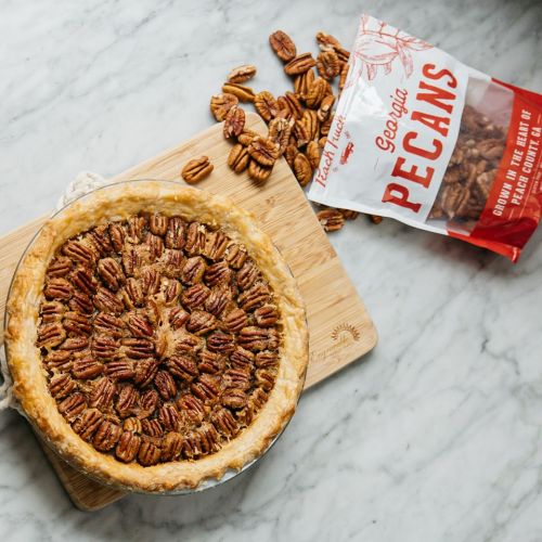 A pecan pie topped with whole pecans, next to a bag of Georgia pecans on a marble surface.