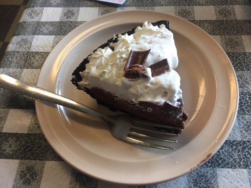 A slice of chocolate cream pie topped with whipped cream and chocolate shavings on a beige plate with a fork.