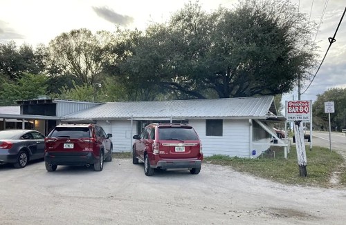 A small barbecue restaurant with a metal roof, surrounded by trees and parked cars in a gravel lot.