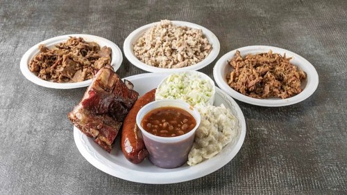 A plate of barbecue featuring ribs, sausage, coleslaw, baked beans, and sides of pulled pork and rice.