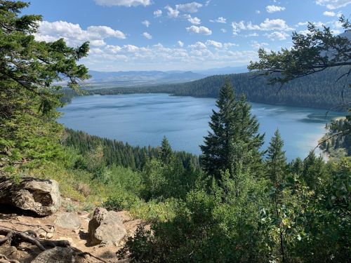A serene lake surrounded by lush green trees and mountains under a blue sky with scattered clouds.