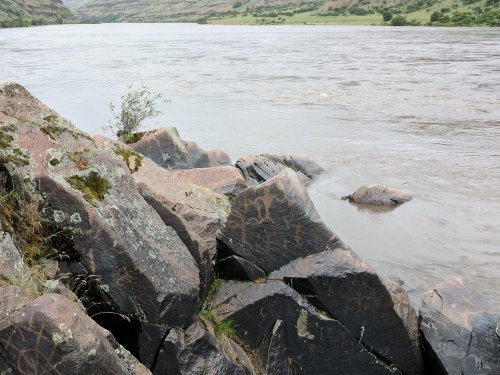 Rugged riverbank with etched rocks and flowing water, surrounded by green hills under a cloudy sky.