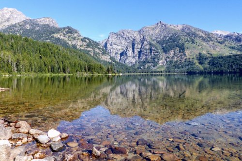 A serene lake reflecting mountains and trees, with clear water revealing stones beneath the surface.