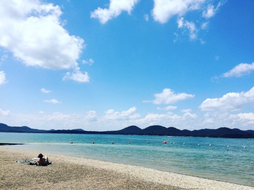 A serene beach scene with a person lounging on the shore, clear blue water, and distant hills under a partly cloudy sky.