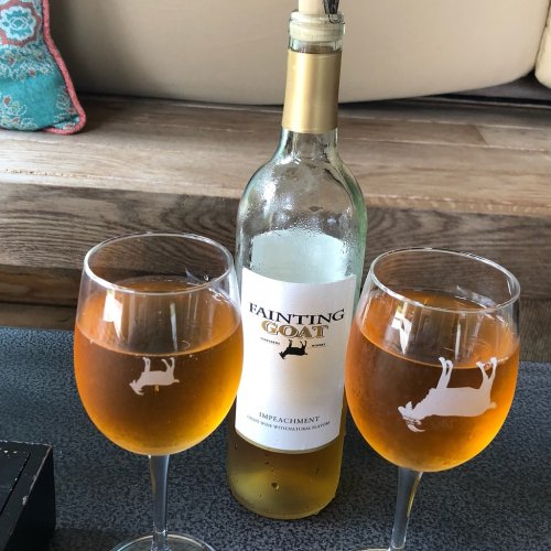 Two glasses of amber wine beside a bottle labeled "Fainting Goat Impeachment" on a wooden table.