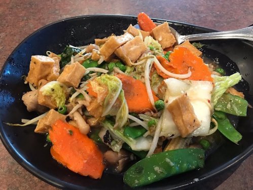 A black bowl filled with mixed vegetables, tofu, and noodles, garnished with bean sprouts and colorful carrots.