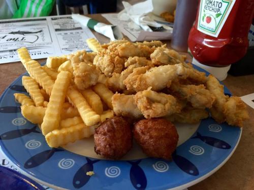 A plate of fried chicken, crinkle-cut fries, and two hushpuppies, with a bottle of ketchup in the background.
