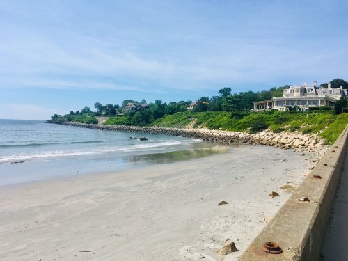 A serene beach scene with a sandy shore, rocky coastline, and lush green hills under a clear blue sky.