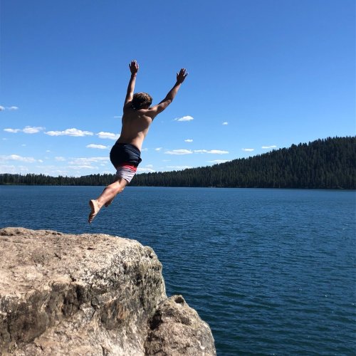 A person jumps off a rock into a lake, surrounded by trees and a clear blue sky.