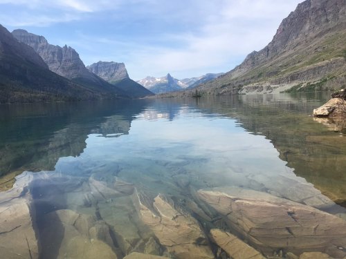 A serene lake reflects mountains and a clear sky, with visible rocks beneath the calm water's surface.