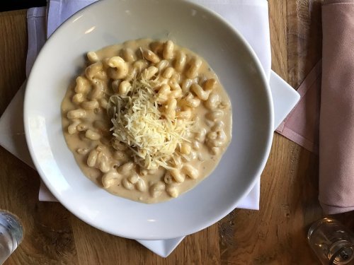 A bowl of creamy macaroni and cheese topped with shredded cheese, placed on a wooden table with napkins and glasses.