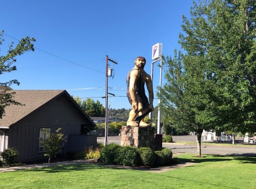 A large statue of a muscular figure stands in a grassy area near a building and a gas station under a clear blue sky.