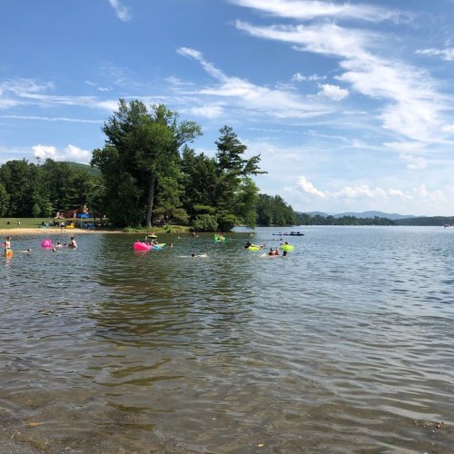 A sunny lake scene with people swimming and floating on colorful inflatables, surrounded by trees and blue skies.