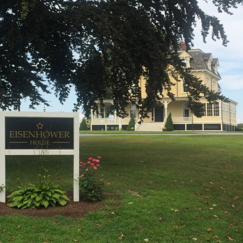 A historic yellow house with a large porch, surrounded by green grass and trees, featuring a sign that reads "Eisenhower House."