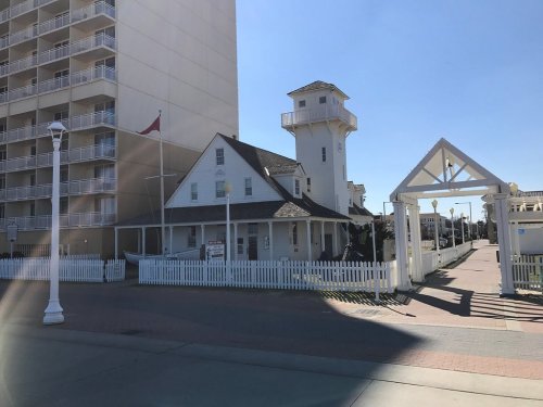 A white building with a tower next to a tall hotel, surrounded by a white picket fence and a clear blue sky.