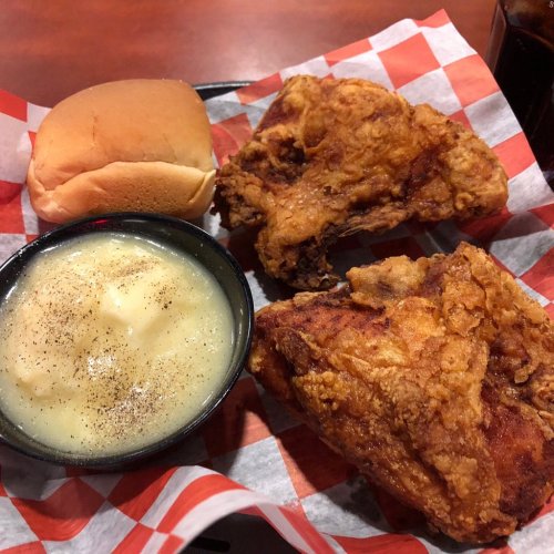 A plate with two pieces of fried chicken, a roll, and a bowl of mashed potatoes with pepper, all on a checkered paper.