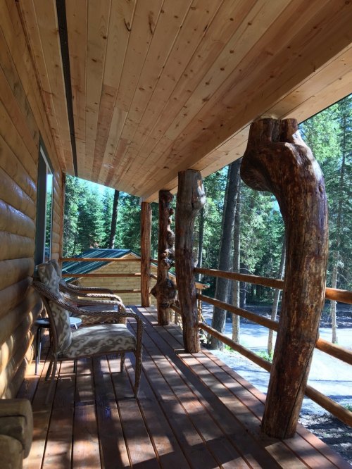 A wooden porch with log pillars, featuring a chair, surrounded by trees and sunlight.