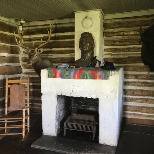 A rustic room with a stone fireplace, a deer head mount, a bust, and a colorful blanket on the mantel.