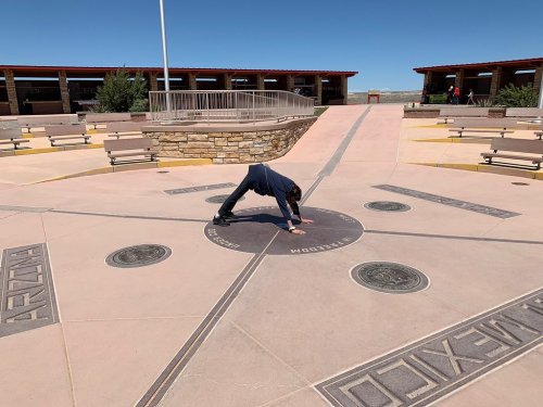A child performs a handstand on a compass rose at a scenic outdoor area with benches and buildings in the background.