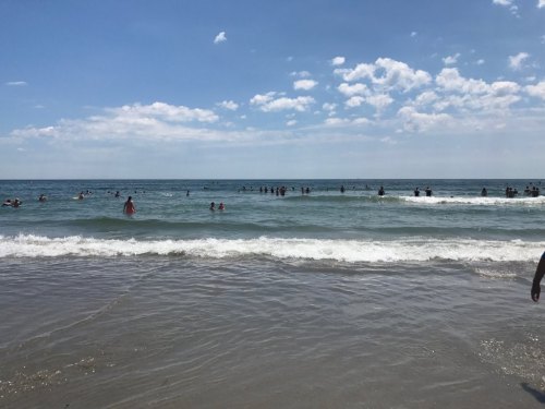 A sunny beach scene with people swimming in the ocean and gentle waves under a blue sky with scattered clouds.