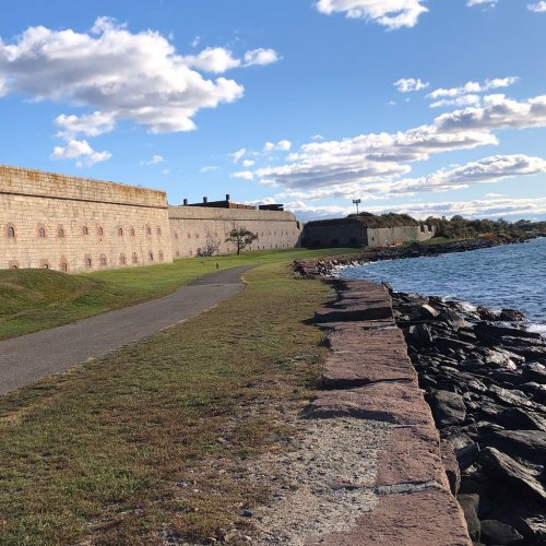 A scenic view of a historic fort along a waterfront, with grassy paths and rocky shoreline under a blue sky.