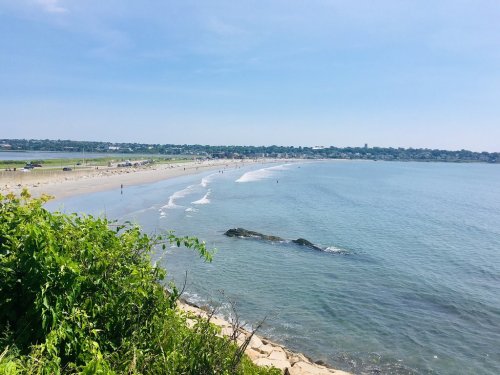 A scenic view of a beach with gentle waves, sandy shore, and greenery in the foreground under a clear blue sky.