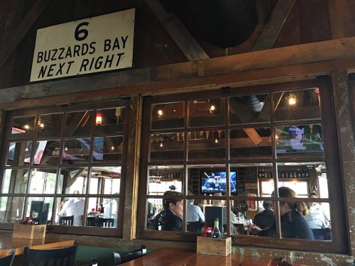 Interior of a rustic restaurant with wooden beams, large windows, and patrons dining. A sign reads "6 Buzzards Bay Next Right."