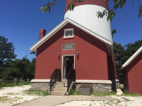 A red lighthouse building with a white top, featuring a plaque and stairs leading to the entrance. Green trees in the background.