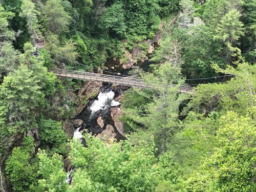 A wooden suspension bridge spans a rocky gorge with a flowing river, surrounded by lush green trees.