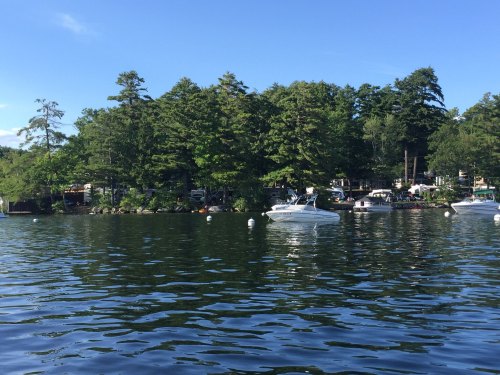 A serene lake scene with boats anchored near a tree-lined shore under a clear blue sky.