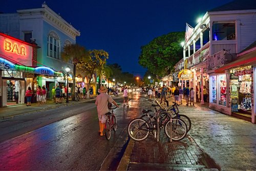 A vibrant street scene at night with shops, bars, and people walking, illuminated by colorful lights. Bicycles parked nearby.