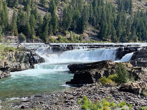 A scenic view of a waterfall cascading over rocky ledges, surrounded by lush green trees and a clear blue sky.