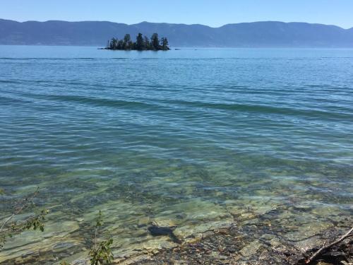A serene lake scene with clear water, rocky shore, and a small island surrounded by mountains in the background.