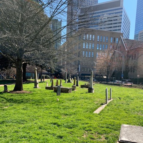 A historic cemetery with gravestones surrounded by grass and modern buildings in the background.