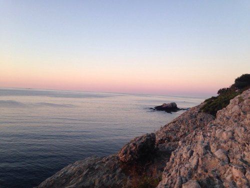A serene coastal scene at sunset, featuring calm waters, rocky shoreline, and a pastel sky.
