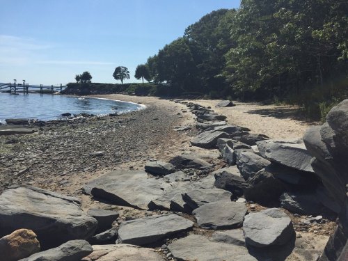 A rocky shoreline with a sandy beach, surrounded by trees and calm water under a clear blue sky.