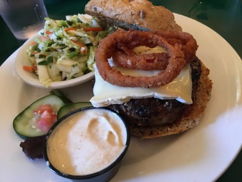 A juicy burger with melted cheese, onion rings, and a side of coleslaw and dipping sauce on a plate.