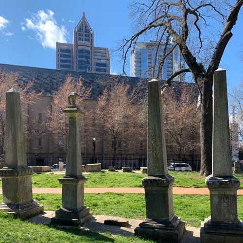 Gravestones in a park with modern buildings and a tree in the background under a clear blue sky.