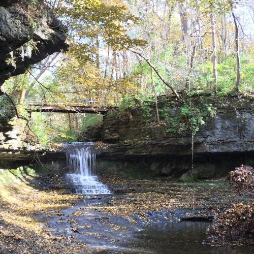 A serene waterfall cascades over rocks, surrounded by autumn foliage and a wooden bridge in a tranquil forest setting.