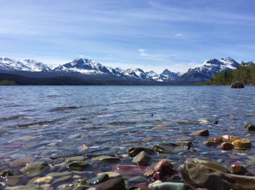 A serene lake with colorful pebbles in the foreground, surrounded by snow-capped mountains under a clear blue sky.