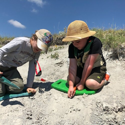 Two children kneel on a sandy surface, focused on digging with tools under a clear blue sky.