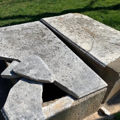 A weathered stone structure with cracked slabs and scattered stones, set on grassy ground.